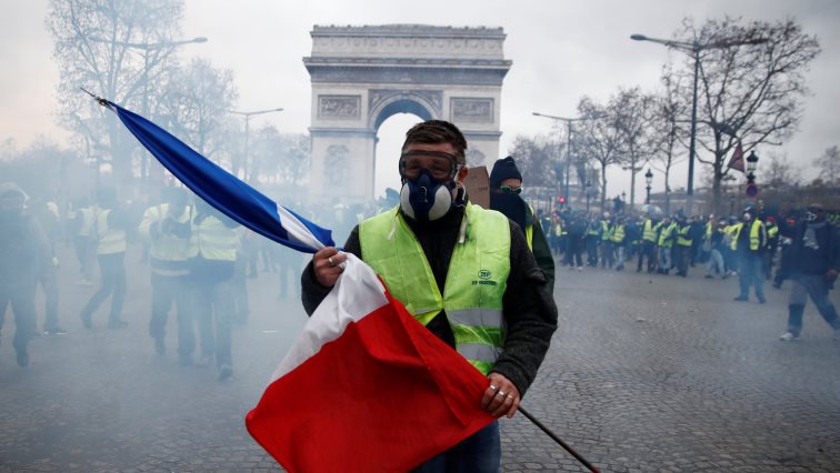 A protester wearing a yellow vest holds a French flag as he walks among tear gas on the Champs-Elysees Avenue