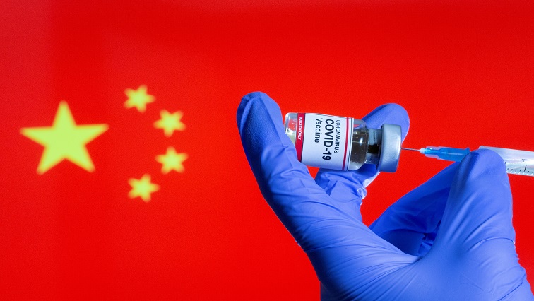 A woman holds a small bottle labeled with a "Coronavirus COVID-19 Vaccine" sticker and a medical syringe in front of displayed China flag.
