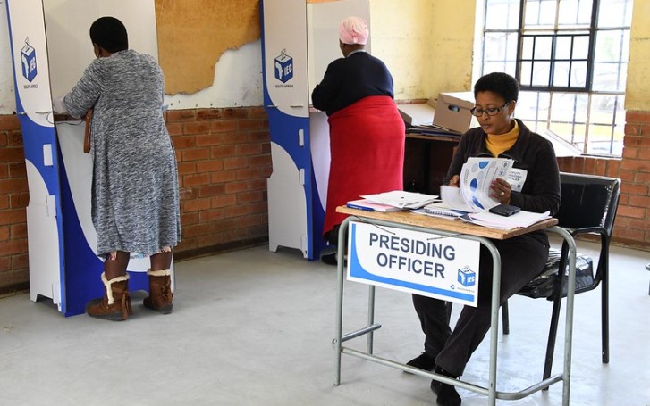 File image | IEC presiding officer sitting at a desk as two people cast their votes in the background.