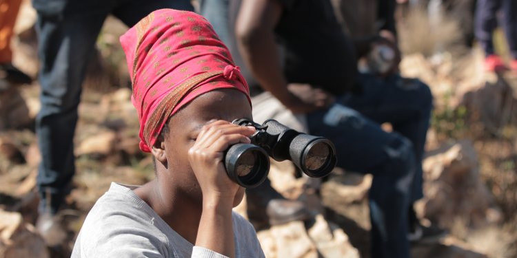 A woman uses binoculars to look into the hills and caves of Munsieville in search of zama zamas said to be hiding in the caves nearby
